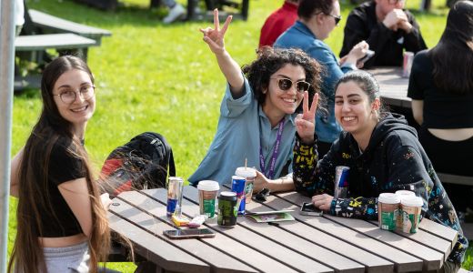 Group of students seated at an outdoor picnic table on grass, with drinks and notebooks, smiling and gesturing towards the camera.