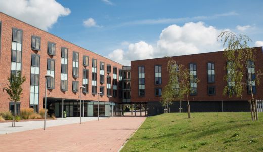 Modern red-brick university building with large windows, a paved courtyard and green lawn under a blue sky.