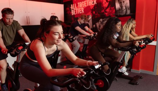 Students using indoor exercise bikes in a gym studio with red lighting.