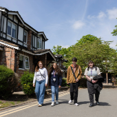 Group of students walking along a tree-lined campus pathway beside residential-style buildings on a sunny day.