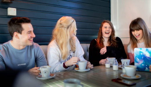 Group of students seated around a café table with cups and saucers, engaged in conversation in a modern indoor setting.