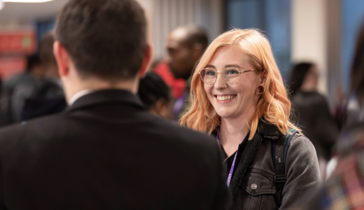 Person wearing glasses smiling while talking at an indoor event, with people and soft lighting in the background.