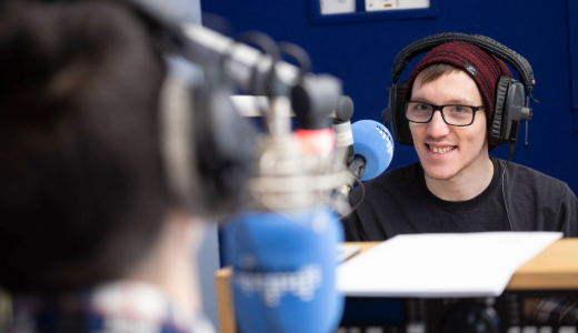 Person wearing headphones speaking into a microphone in a radio studio, with recording equipment and blurred foreground.