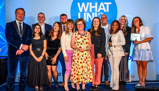 Group of award recipients standing on a stage holding certificates, with a “WHAT UNI?” backdrop behind them.