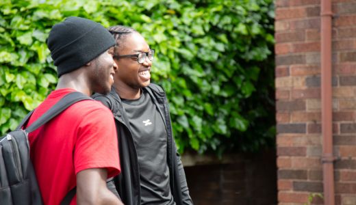 Two people walking and smiling beside a brick building, with green foliage in the background; one carries a backpack.