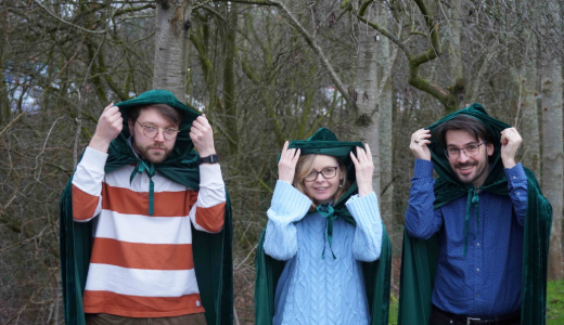 Three people standing outdoors in a wooded area, each wearing a green cloak and lifting the hood over their head.