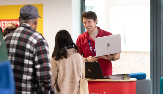 An information desk representative holding a laptop speaks with visitors at an indoor event.