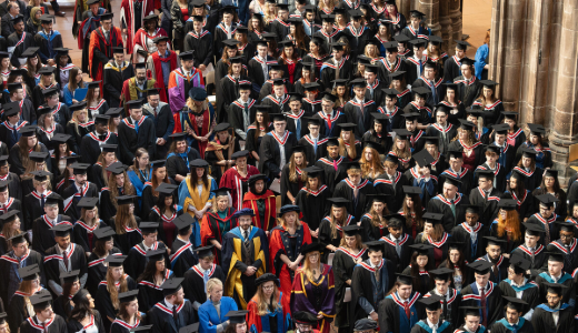 A large group of graduates in caps and gowns seated and standing in a grand hall during a graduation ceremony, with academic staff in colourful robes positioned among them.