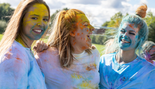 Three people outdoors wearing white T‑shirts covered in brightly coloured powder, standing close together during a colour‑run style event.