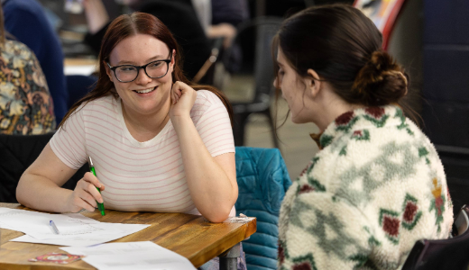 Two students sitting at a table discussing paperwork together in a busy indoor environment