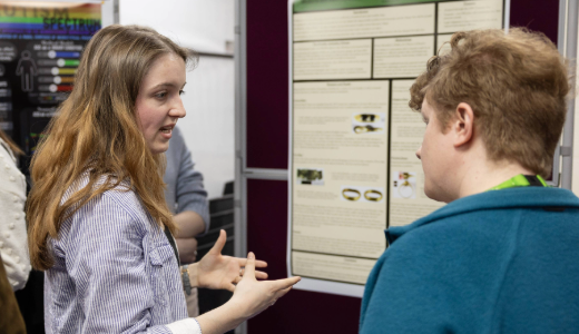 Two people stand in front of an academic poster, with one explaining the content.