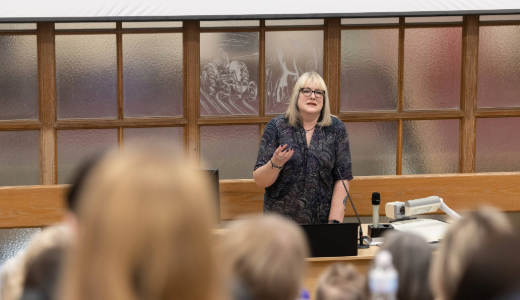 A presenter stands at the front of a lecture room, speaking to an audience.