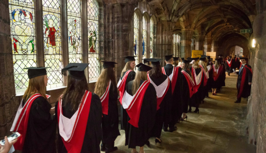 Graduates in caps and gowns walk in a line through Chester Cathedral.