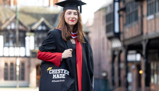 A graduate in cap and gown stands outside historic black‑and‑white buildings, holding a tote bag that reads ‘Chester Made’.