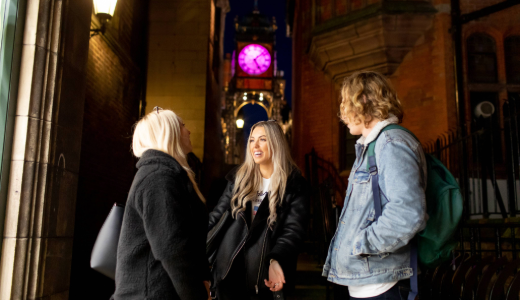 Three people talk together in a narrow street at night, with a brightly lit clock tower glowing pink in the background.