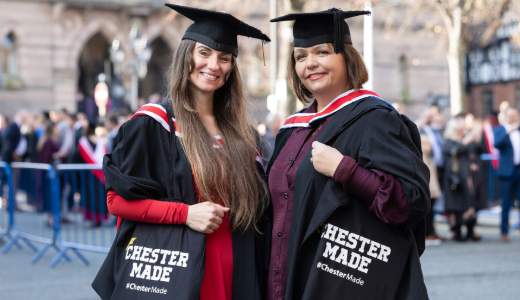 Two graduates in academic gowns and caps holding “Chester Made” tote bags outside on graduation day.