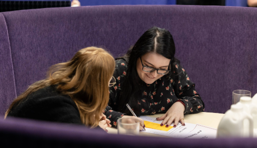 Two people seated in a purple booth, reviewing and writing on paperwork spread out on the table.