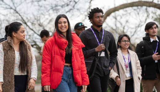 A group of students walking across campus, some wearing lanyards, with trees in the background.