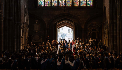 A graduation ceremony inside a cathedral, with graduates in gowns and an audience seated beneath stained‑glass windows.