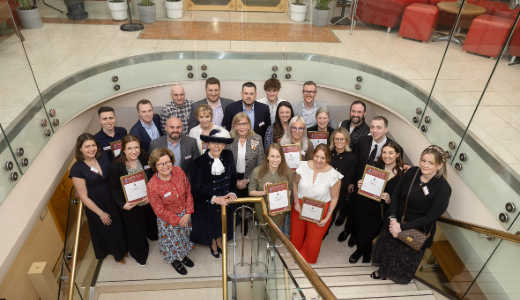 A large group gathered on a staircase indoors, several holding framed certificates.
