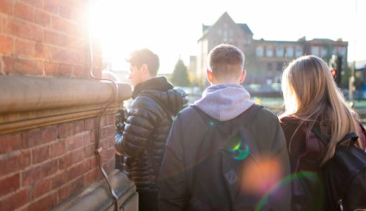 A group of three people walking beside a brick building on a sunny day, with light flaring into the lens.