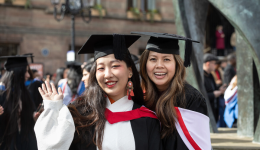 Two graduates wearing academic gowns and mortarboards, standing outdoors during a graduation event.