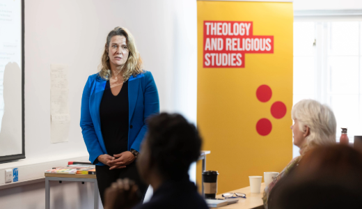 A speaker standing at the front of a classroom, with a banner reading “Theology and Religious Studies” behind them.