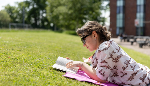 A person lying on grass reading a book near campus buildings on a sunny day.