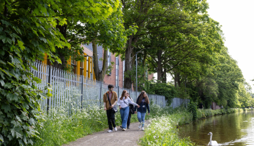 Three people walking along a tree-lined path beside a canal, with greenery and a swan visible in the water.