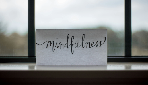 Card with the word “mindfulness” written in cursive, placed on a windowsill with a softly blurred outdoor background.
