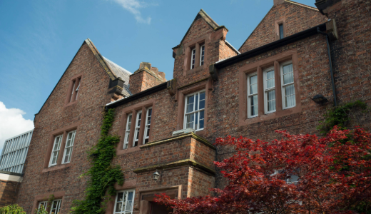 Red-brick historic university building with gabled roofs, large windows, and surrounding foliage.