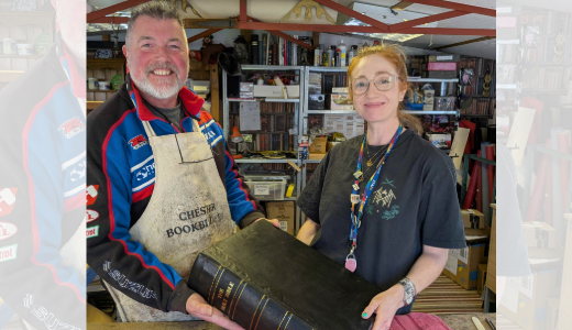 Two people inside a workshop holding a large bound book, surrounded by tools and shelves of materials.