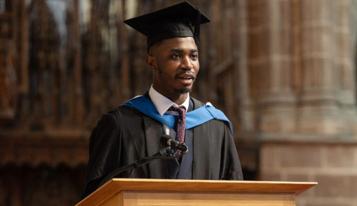 Graduate in academic dress speaking at a lectern inside a historic hall during a ceremony.