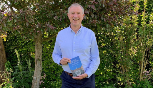 Person standing outdoors in a garden, holding a book and posing in front of greenery.