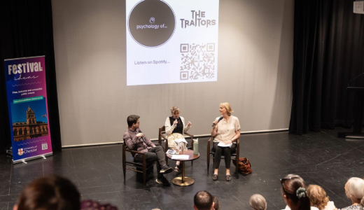 Panel discussion on stage with three speakers seated and a projected screen behind displaying “The Traitors” and a QR code.