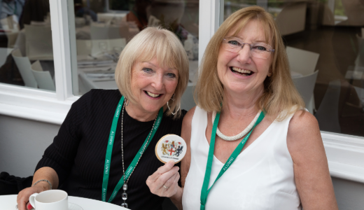 Two attendees wearing green lanyards sit together at an event table, with one holding a commemorative badge.