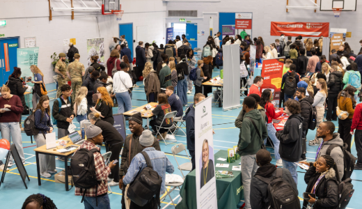 Crowded recruitment fair in a sports hall, with attendees speaking to exhibitors and visiting information stands.