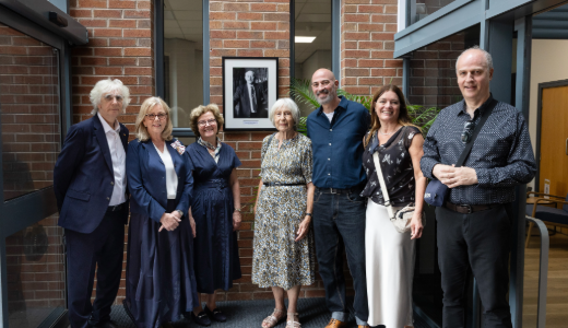 Group photo of six people standing in a university building foyer with a framed portrait displayed on a brick wall behind them.