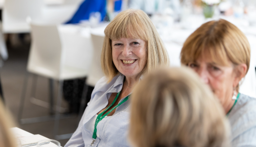 Person wearing a lanyard seated at a table during a social event, with other attendees visible in the foreground and background.