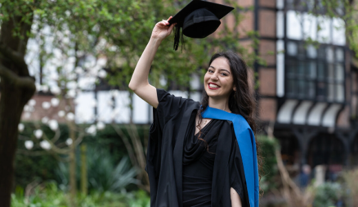 Graduate in academic gown and cap standing outdoors, holding a mortarboard aloft in a garden setting.
