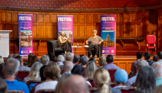 Audience attending a festival talk in a wood-panelled hall, with speakers seated on stage and event banners displayed behind them.