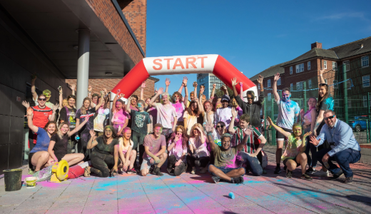 Group of participants covered in coloured powder posing beneath a start arch at an outdoor run.
