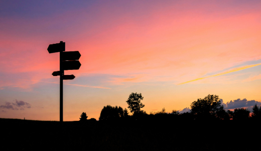 Silhouetted signpost against a colourful sunset sky with trees on the horizon.